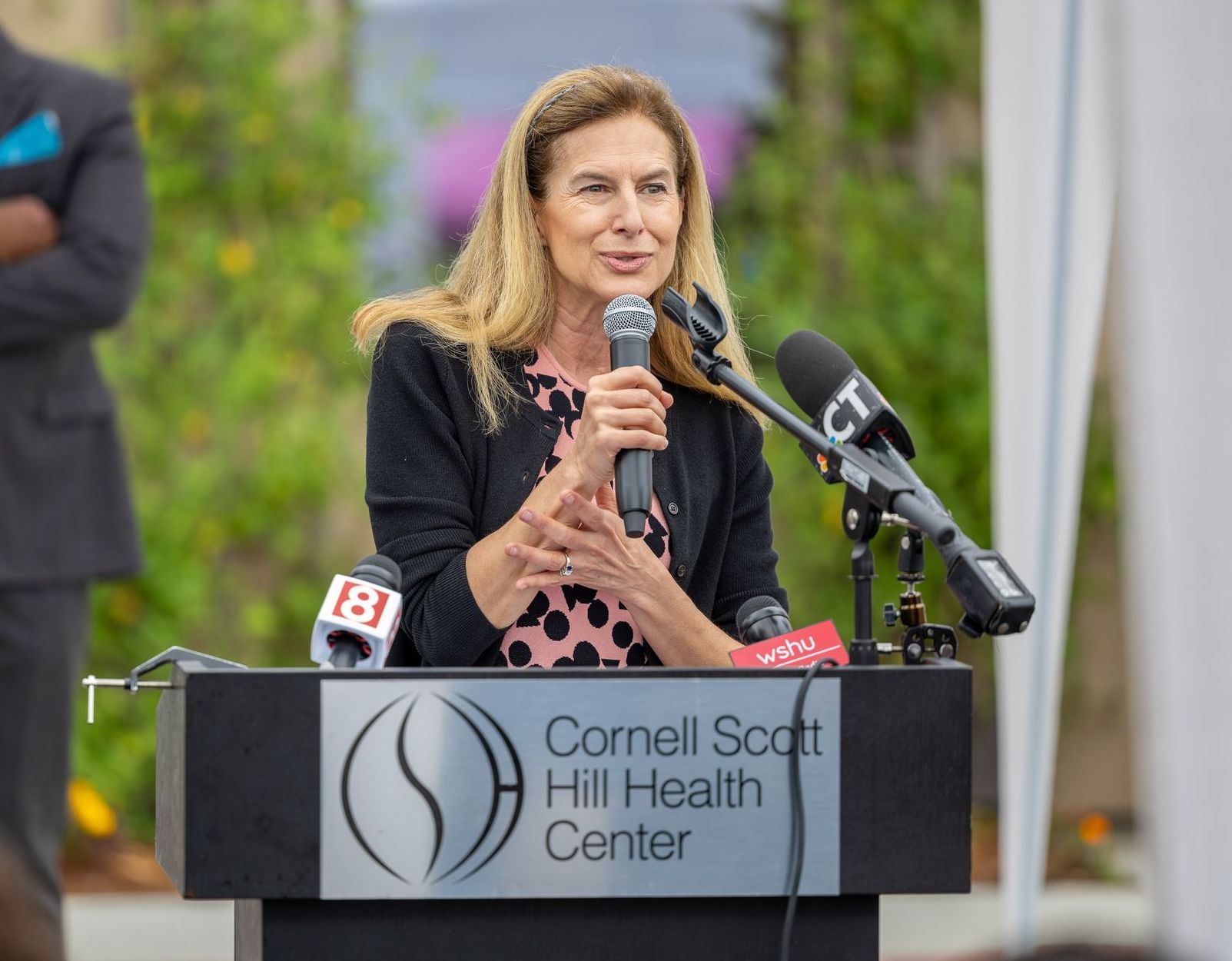 Speaker at podium at Cornell Scott Hill Health Center, holding a microphone during an outdoor event.