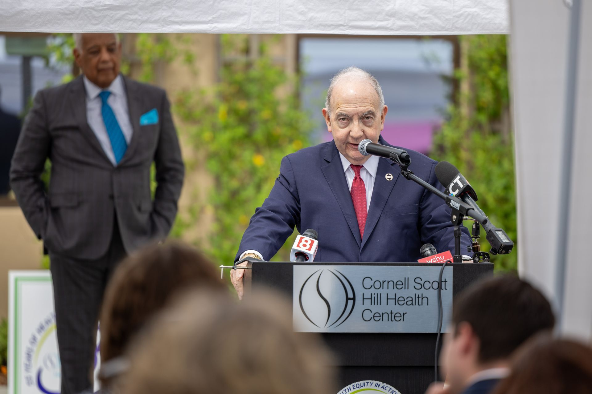 Man speaking at Cornell Scott-Hill Health Center podium during an outdoor event, with an audience in front