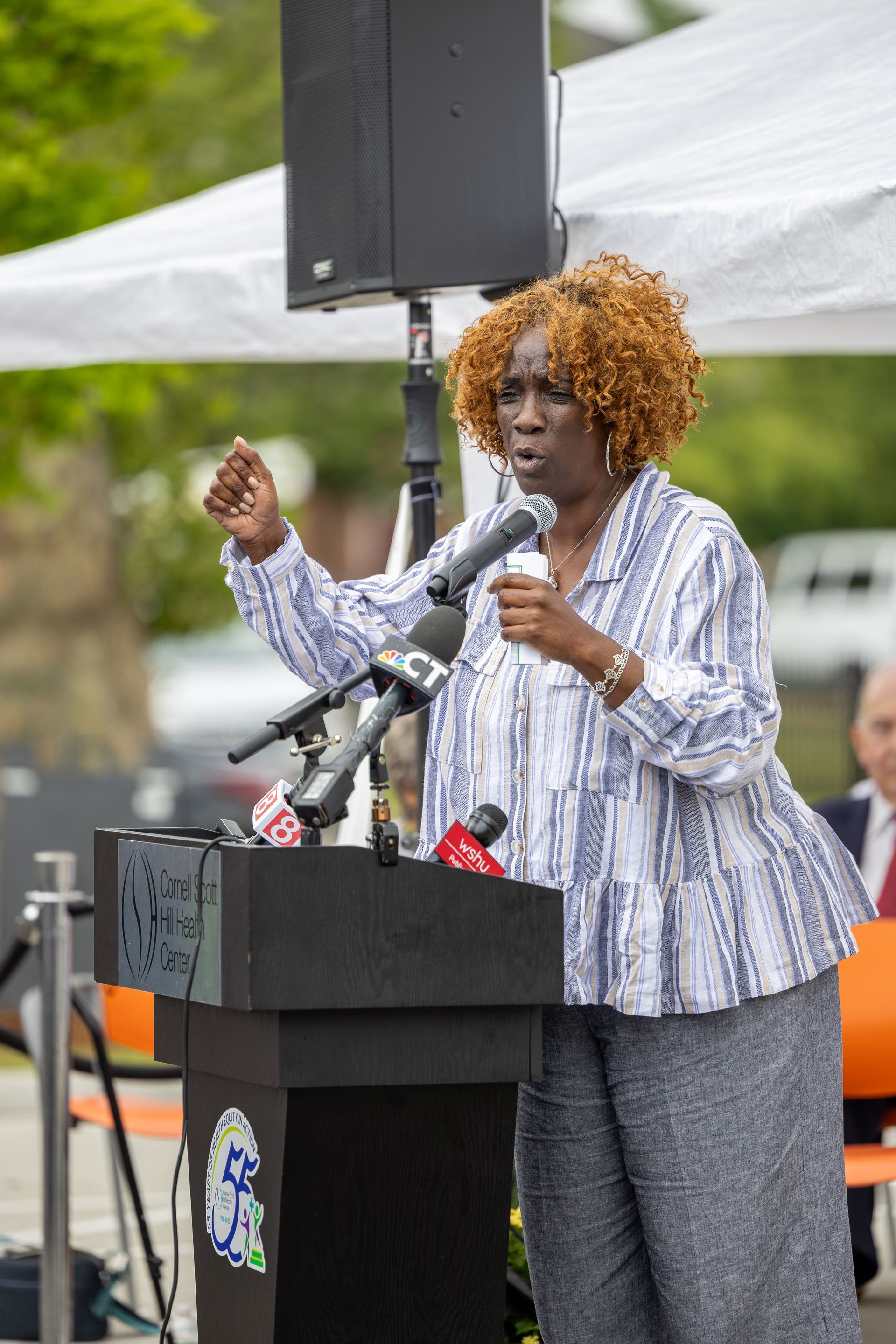 Speaker at podium gesturing into multiple microphones outdoors under a white canopy.