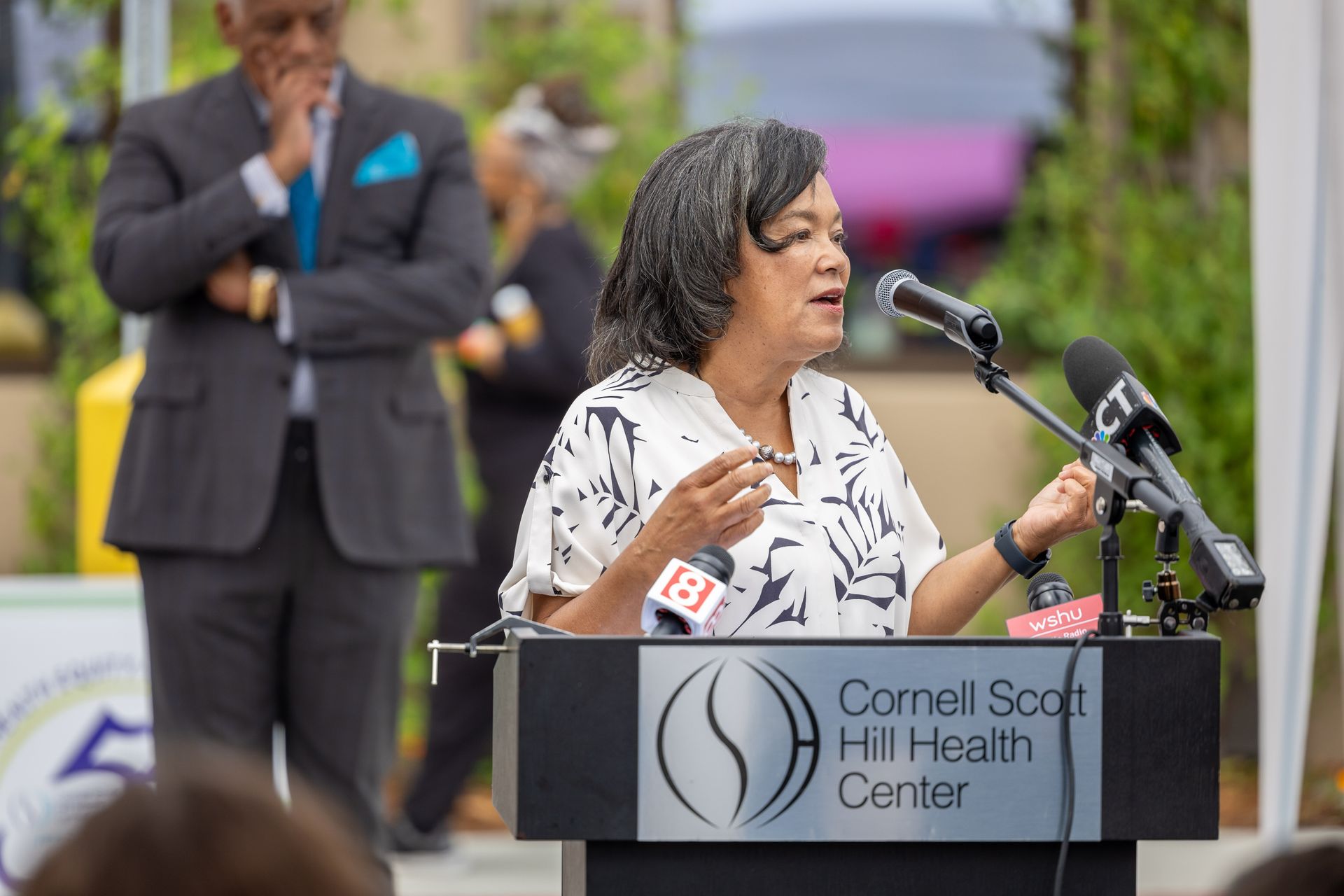 Speaker at Cornell Scott-Hill Health Center podium addressing an outdoor audience, with microphones and a blurred man behind.
