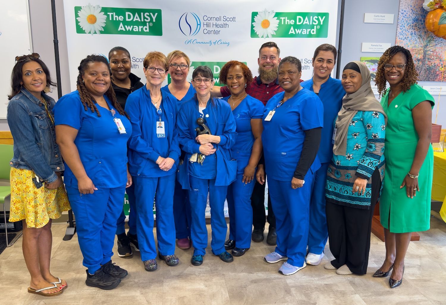 Group of smiling staff in blue and green uniforms posing in front of The DAISY Award banner
