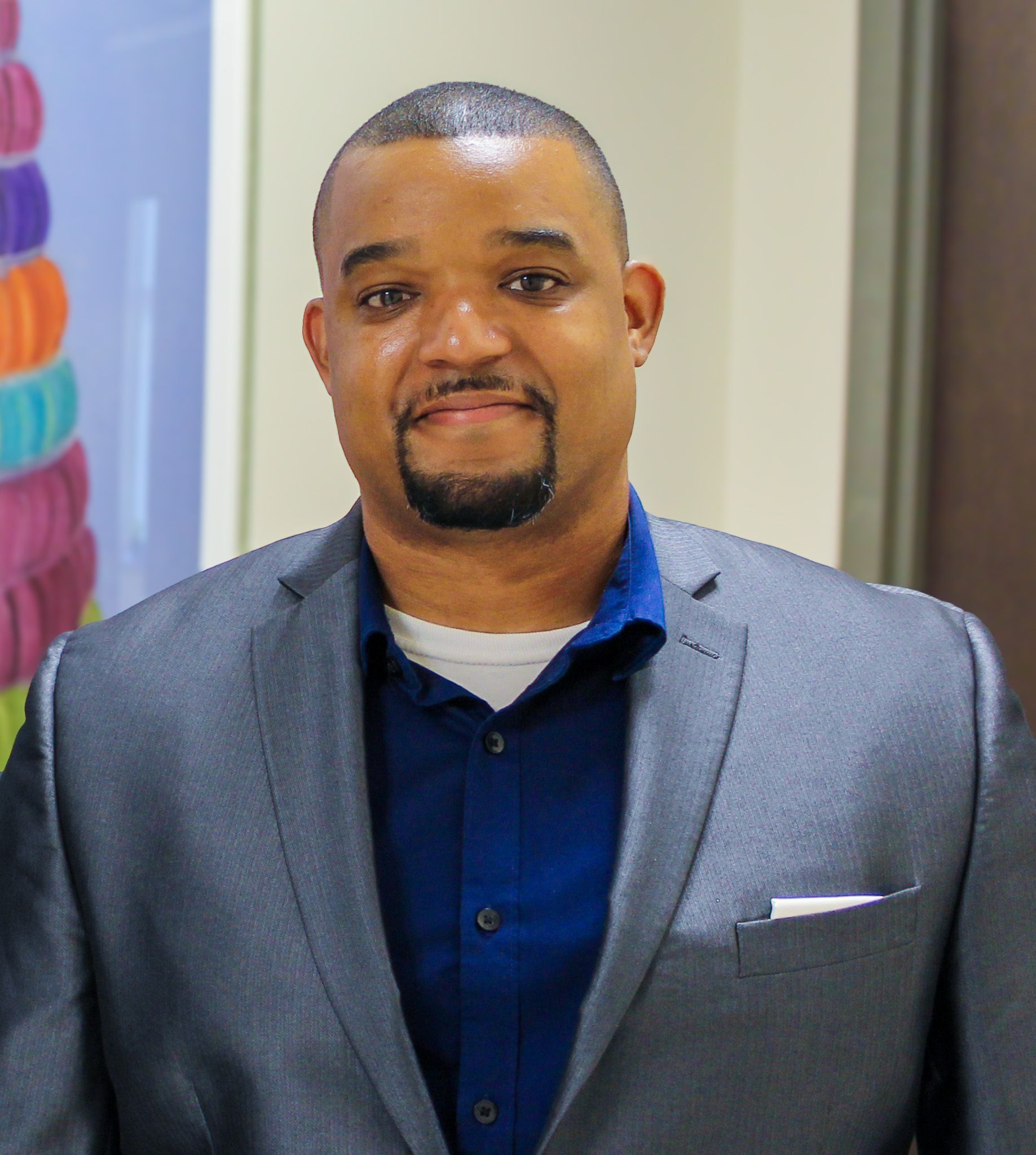 Man in a gray blazer and blue shirt standing indoors, looking at the camera with a slight smile