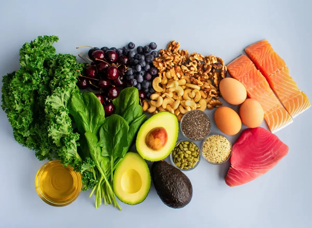 Assorted healthy foods on a light surface: vegetables, fruits, nuts, salmon, eggs, seeds, and avocado.