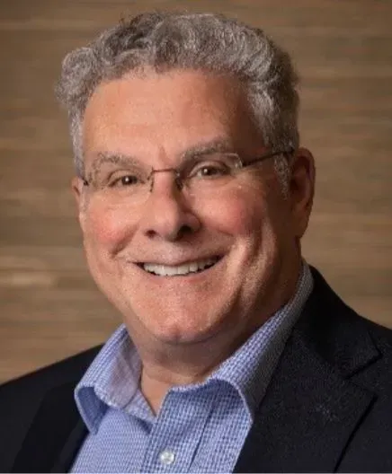Portrait of a smiling man in glasses, wearing a navy blazer and blue shirt against a warm brown background