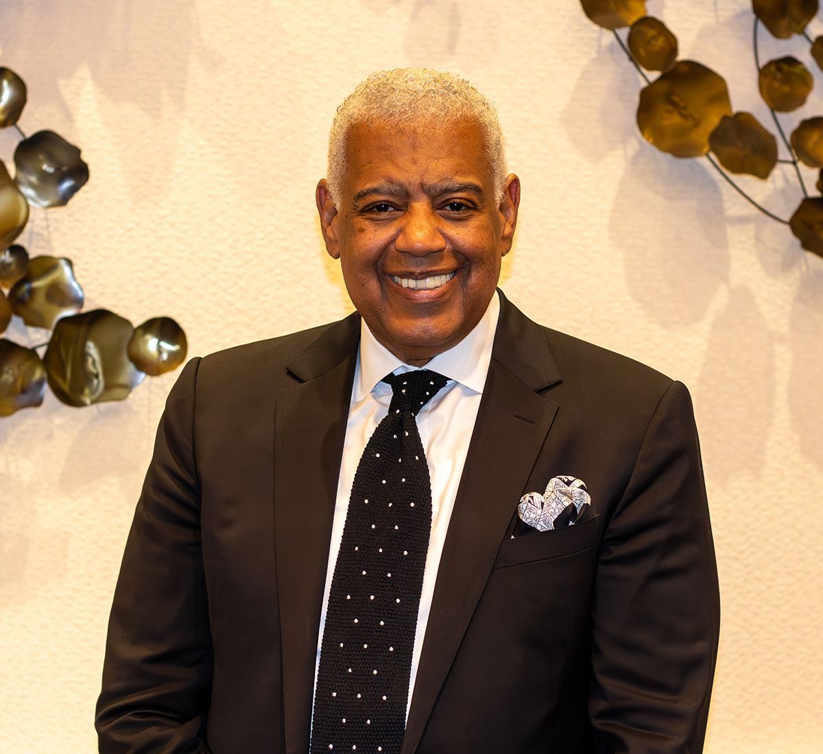 Smiling man in a dark suit and tie standing indoors against a light wall with decorative leaves