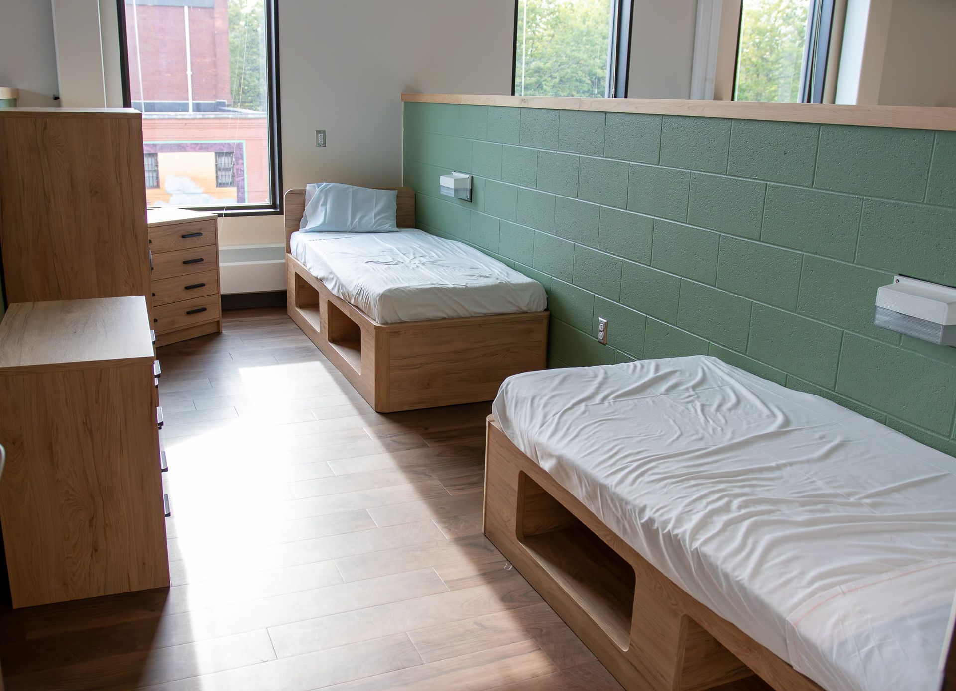 Small dorm room with two single beds, wooden furniture, and a green accent wall by a window.