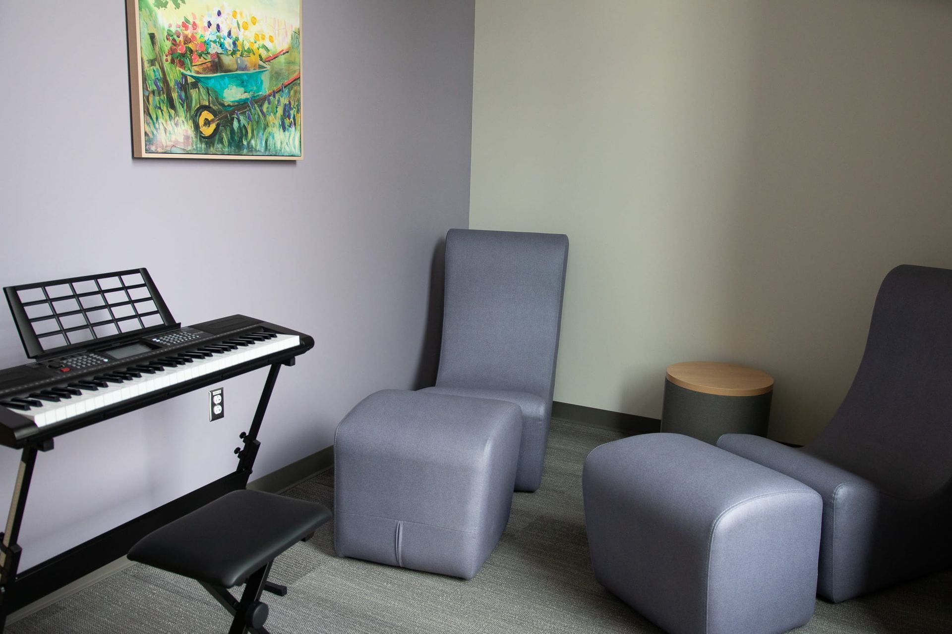 Small music room with keyboard, stool, gray lounge chairs, and colorful wall art.
