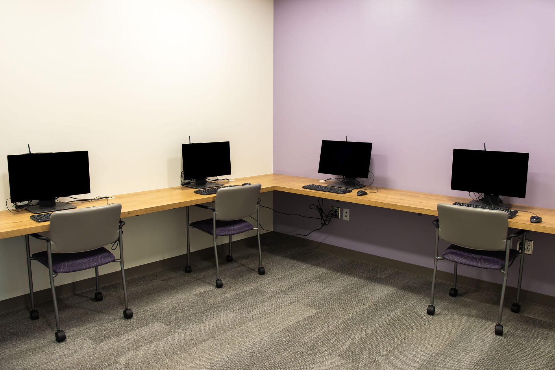 Empty computer lab with four desktop monitors on wooden desks and gray chairs against beige and lavender walls