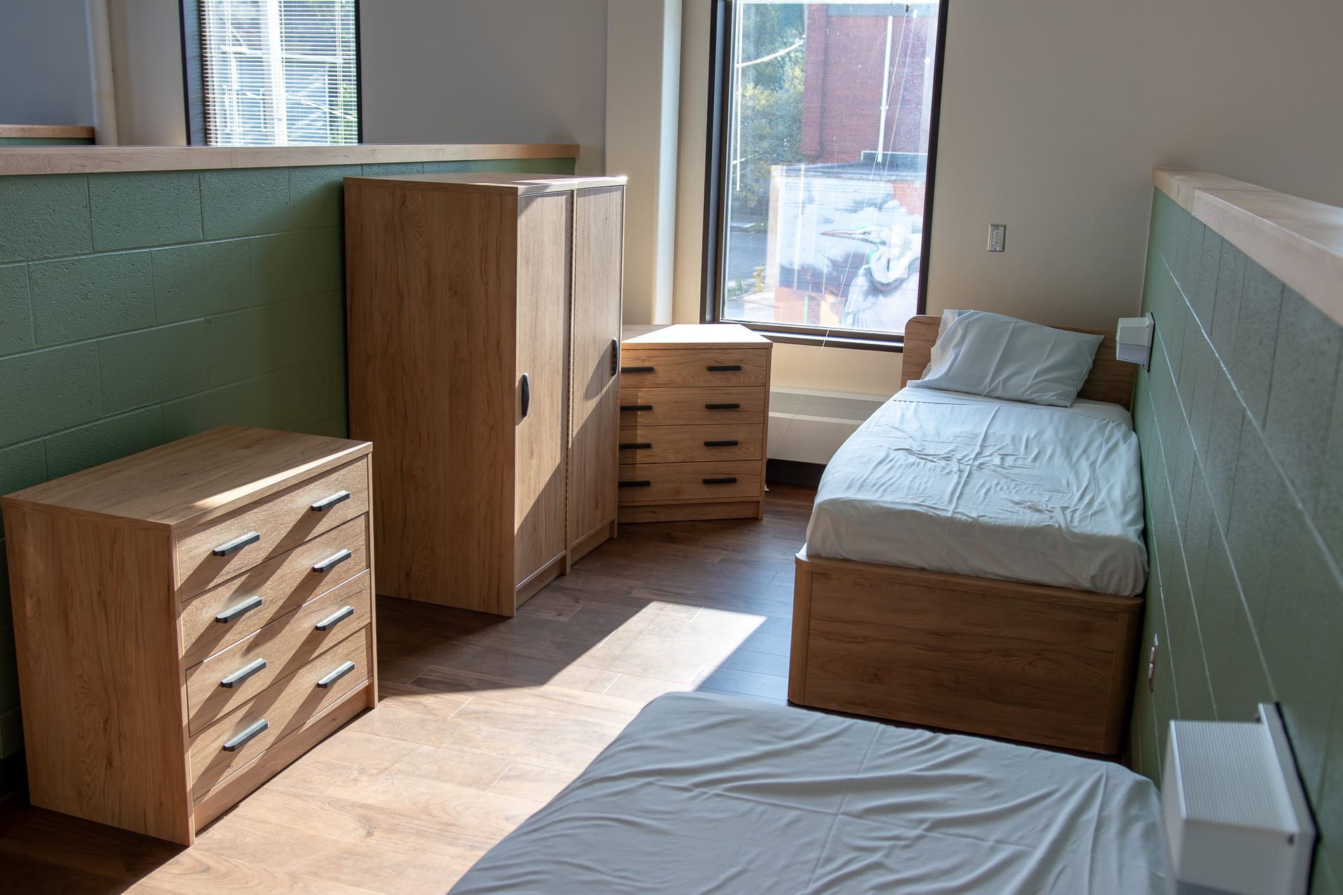 Small dorm room with two beds, wooden dressers, and a window letting in sunlight.
