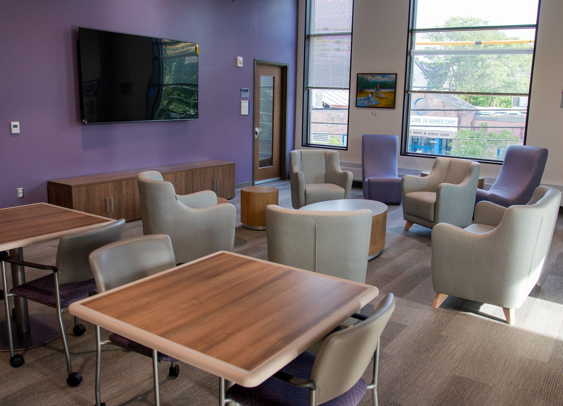 Modern lounge with purple walls, TVs, and gray chairs around wooden tables by large windows
