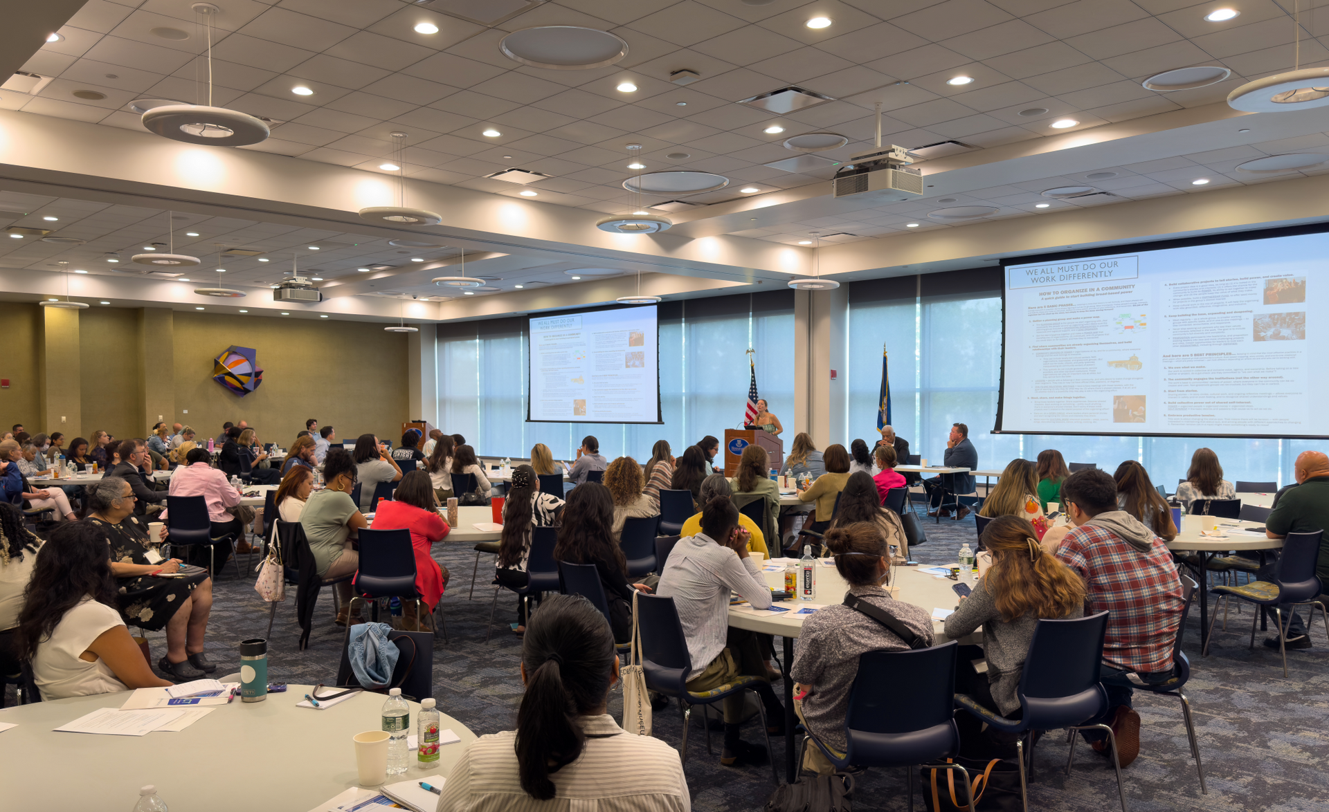 A large conference room with tables and chairs. People are listening to a speaker at the podium