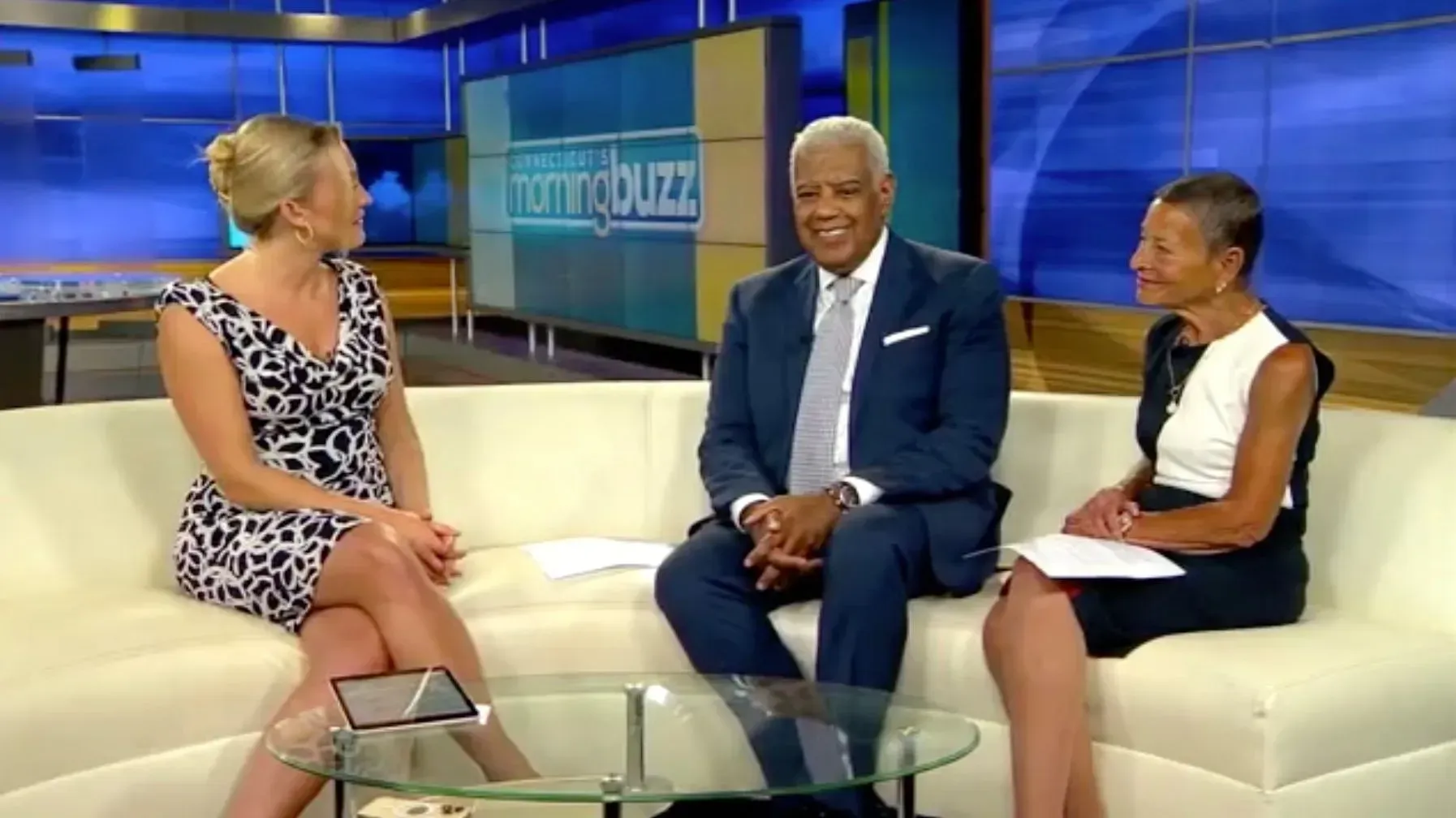 Three people seated on a white couch in a TV studio set, talking during a news interview.