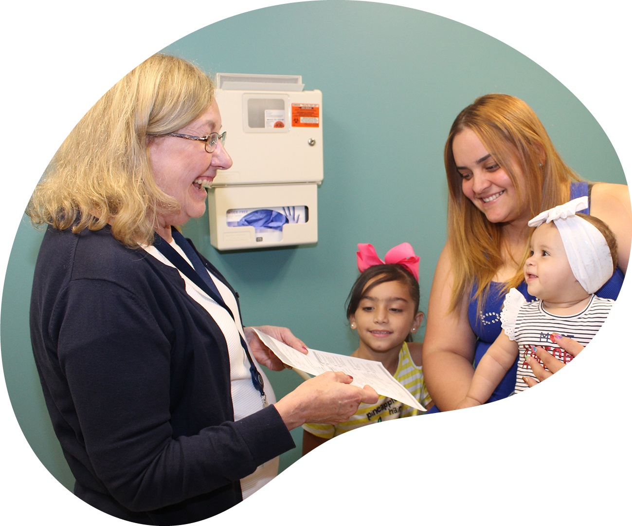 Woman at a clinic smiling with a mother and two children, one holding a baby, in a waiting room.