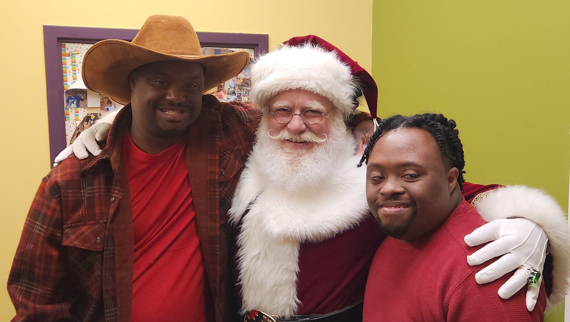 Two men pose with Santa Claus indoors. One man wears a cowboy hat, the other has dreadlocks. All smile.