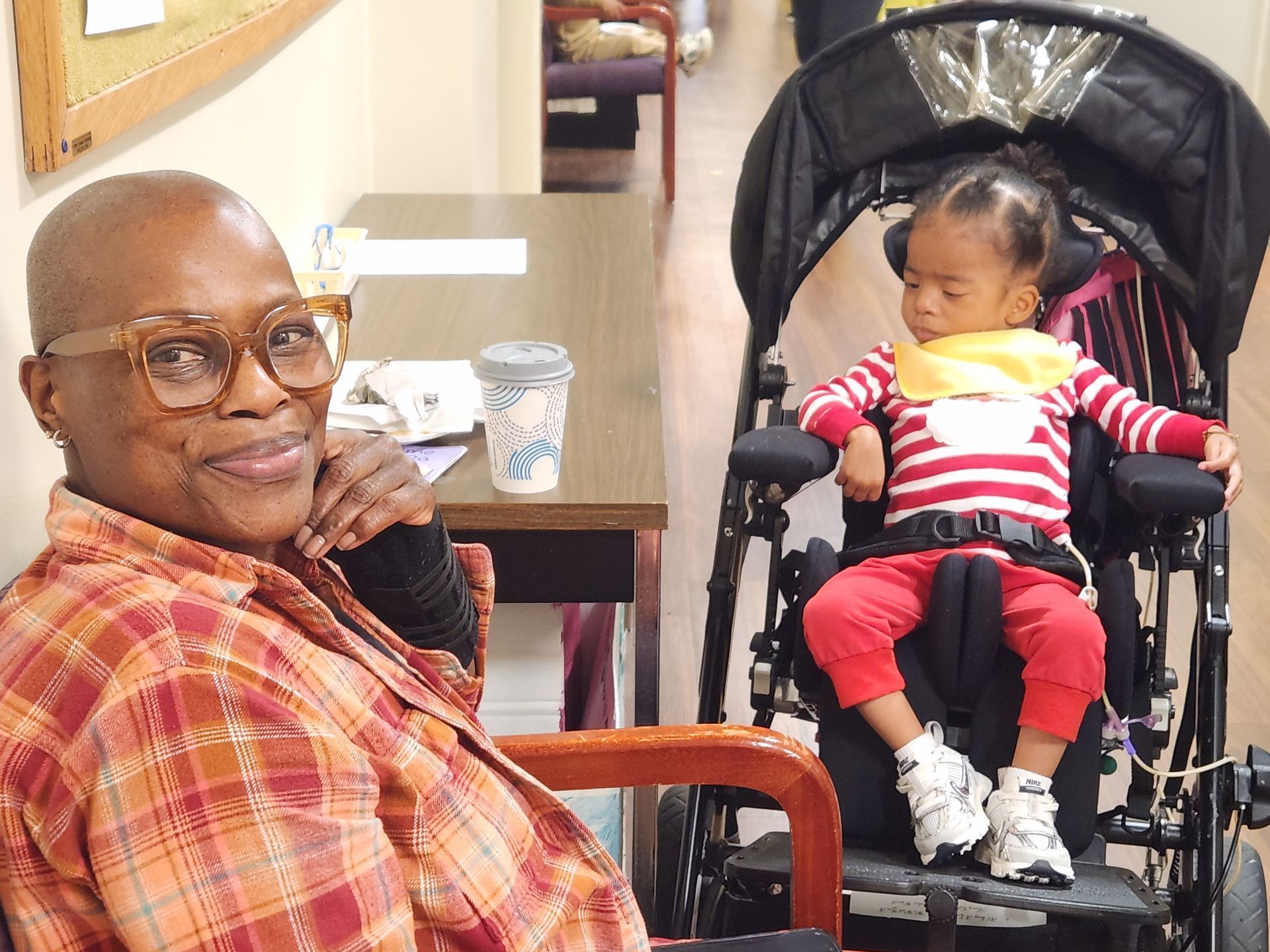 Woman in orange plaid shirt smiles next to child in stroller, wearing red pants and white shoes. Indoor setting.