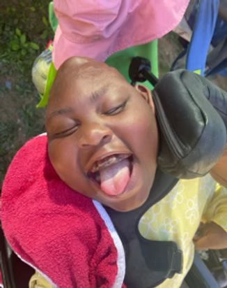 Smiling person with head support, tongue out, wearing a pink hat and yellow shirt.