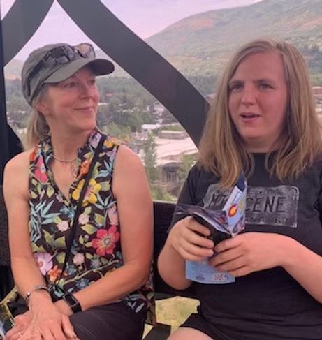 Two women in a gondola, one with a floral shirt, the other holding a map. Mountains visible outside.
