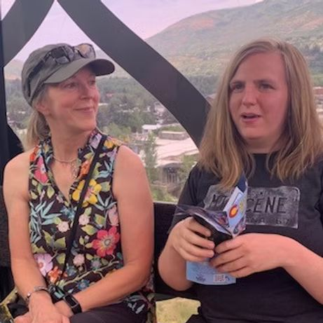 Two people sit inside a gondola, one with a floral top and hat, the other with a graphic tee holding papers.
