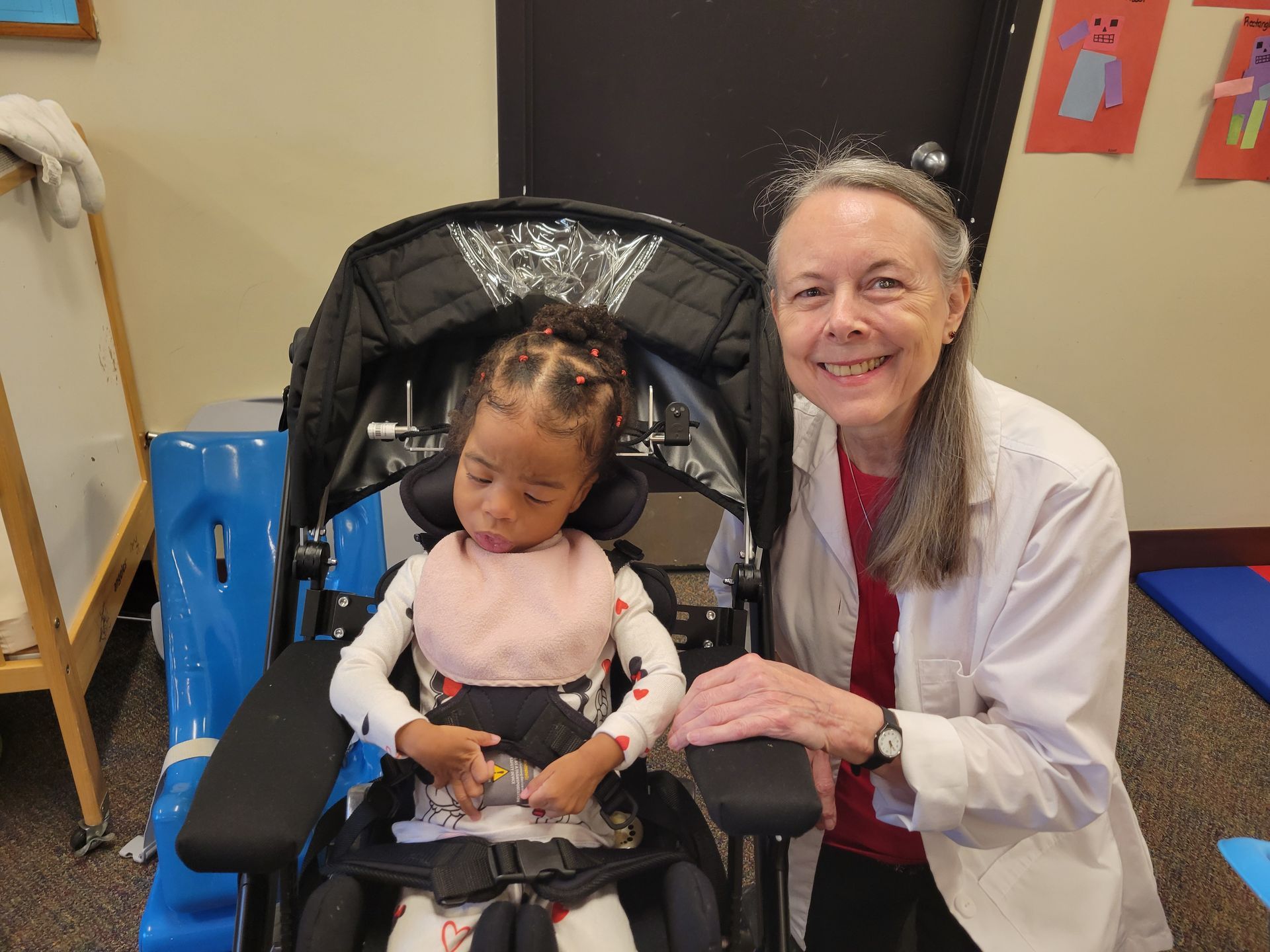 Woman with child in a wheelchair. Both smile, indoors. Child wears a bib, woman has a lab coat.