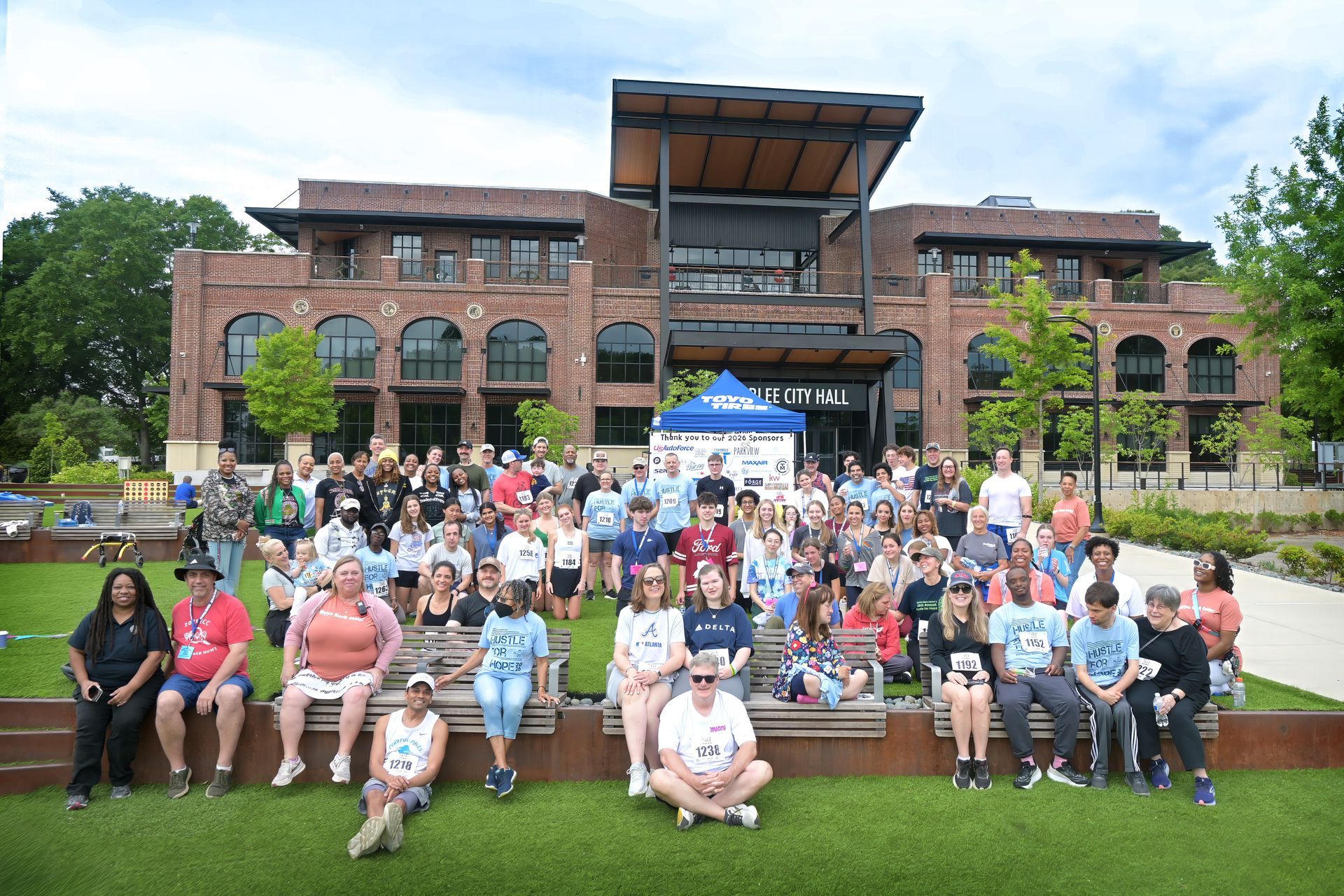 Group of people pose on grass with building backdrop, some wearing race bibs, event.