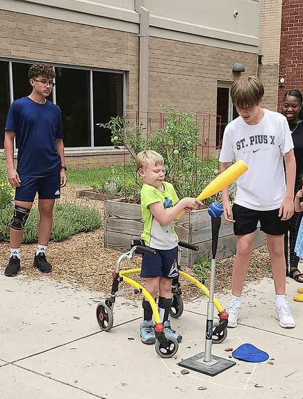 Boy using a walker bats at a tee ball. Two other boys and a woman watch outdoors.