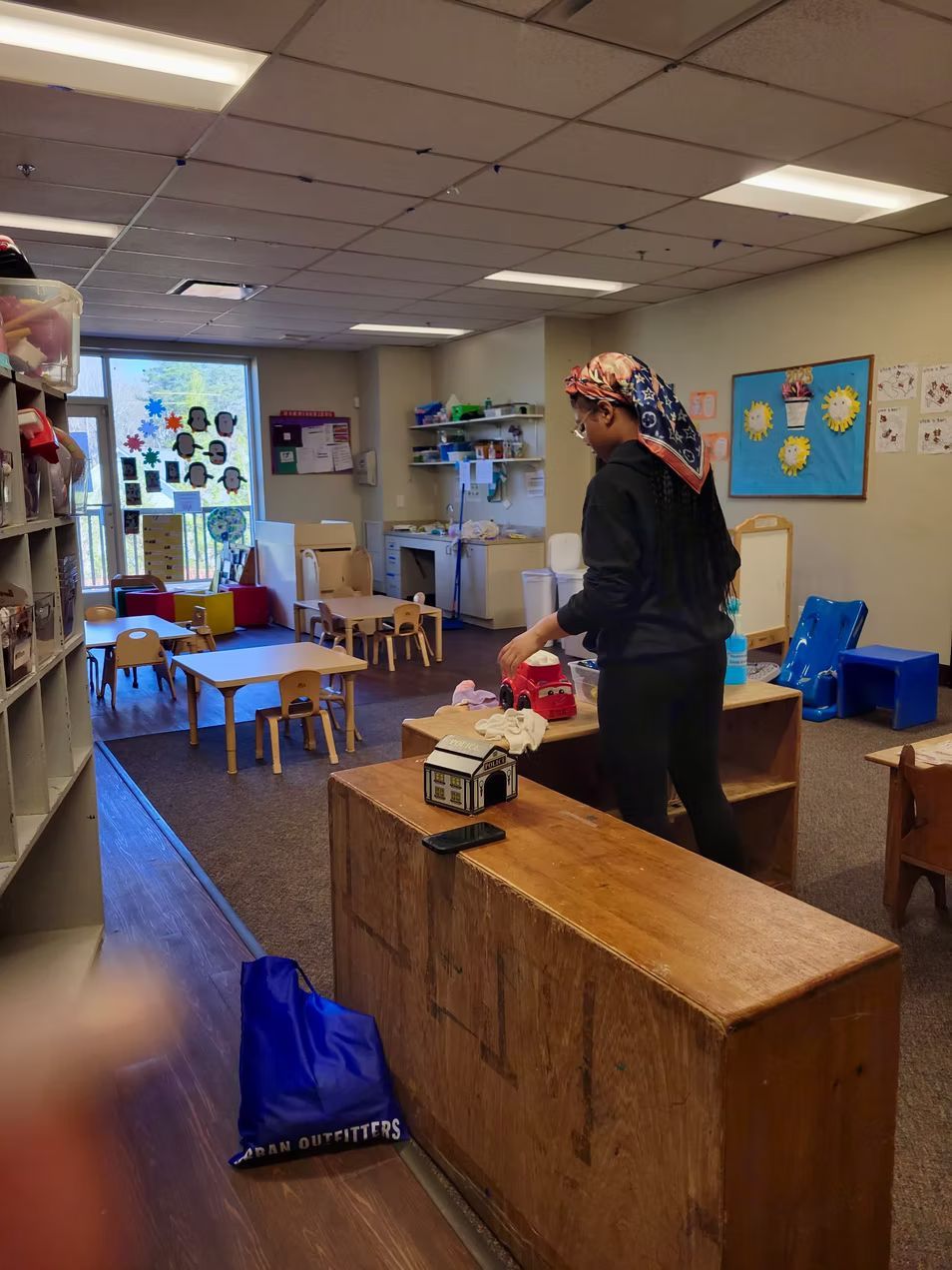 A person in a daycare setting puts away items. Tables, shelves, and play areas are visible.