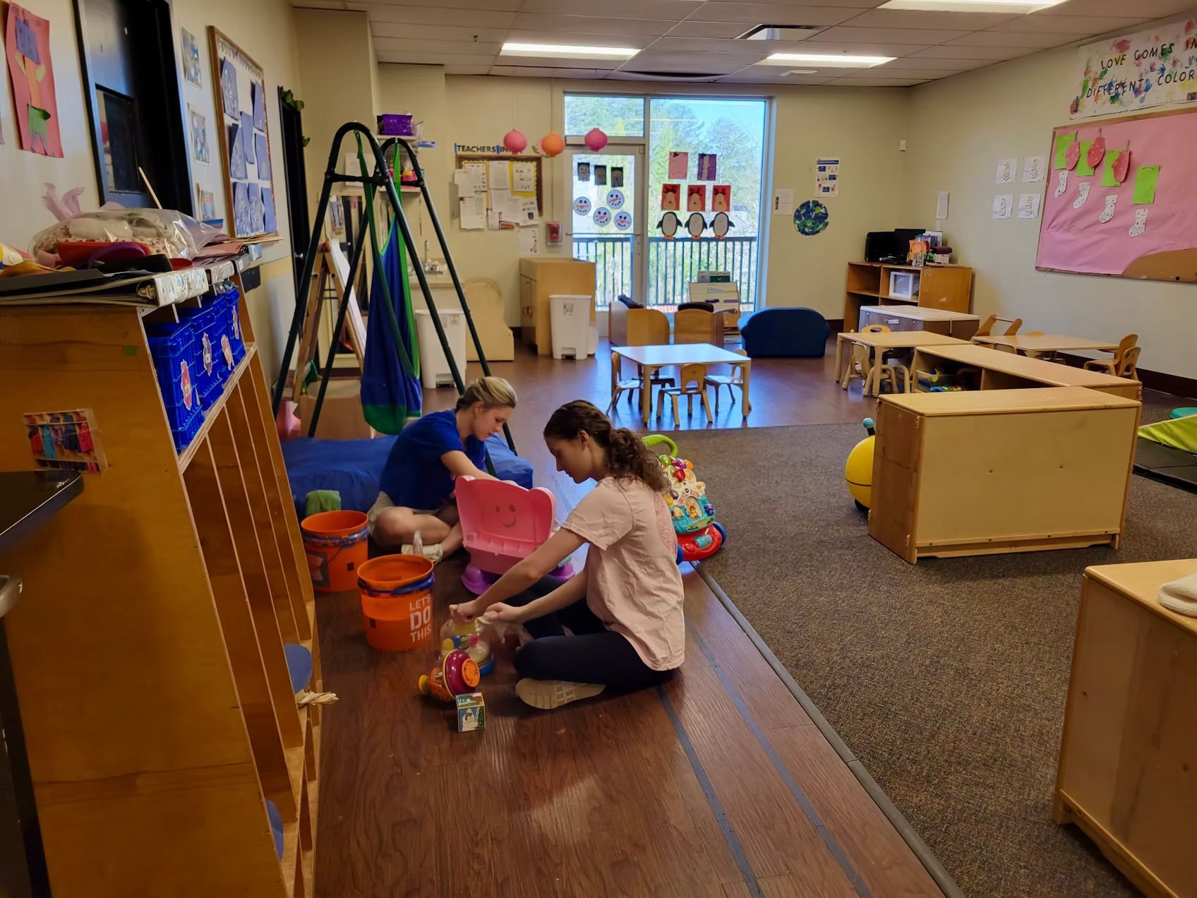 Two people on the floor in a brightly lit playroom arranging toys. Wooden shelves line the left wall, and a doorway is in the center.