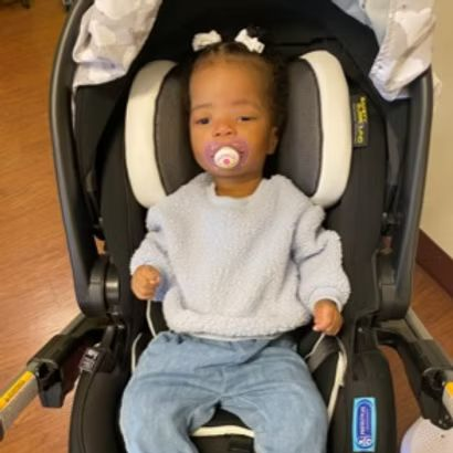 Baby in a car seat, wearing a blue sweater and jeans, with a pacifier.