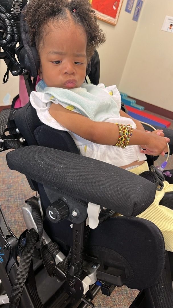 Child in wheelchair with armrests, wearing headphones and bracelet, in a room.