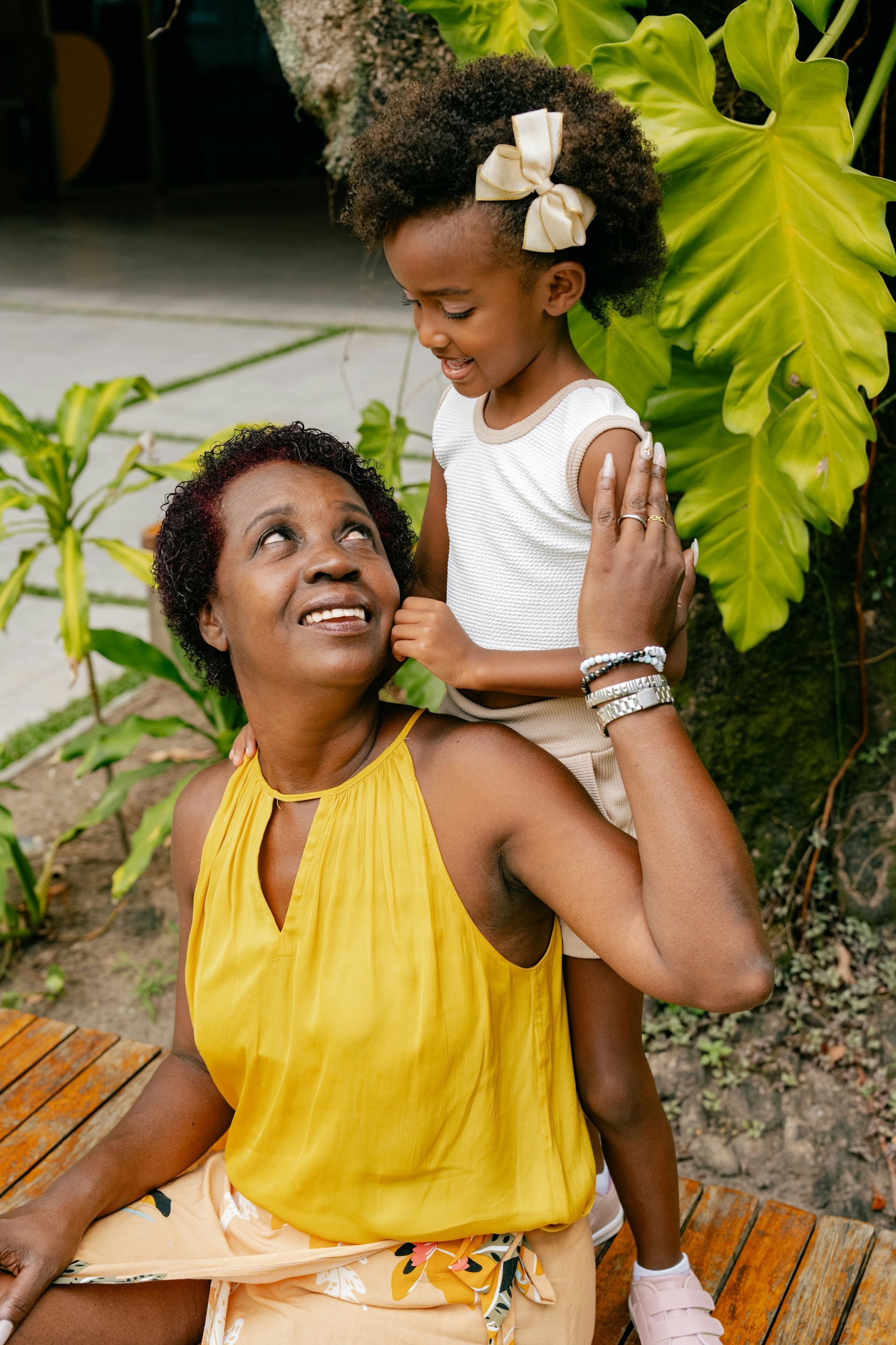 A woman is sitting on a wooden bench holding a little girl on her shoulders.