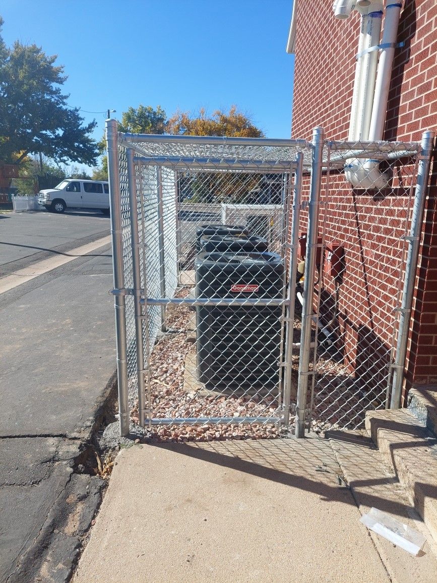 Chain-link cage around outdoor utility equipment beside a brick building and sidewalk.