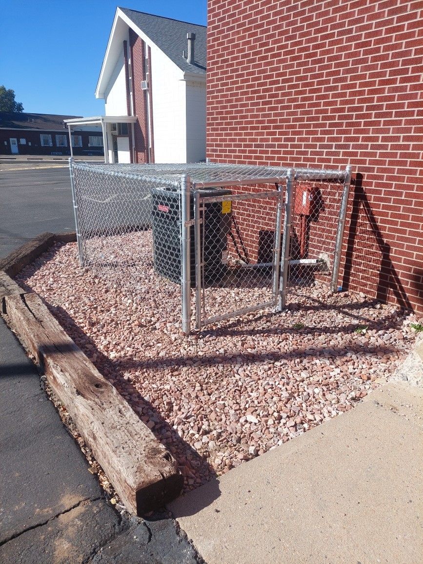 Chain-link utility enclosure beside a red brick building, with gravel ground and a wooden barrier.
