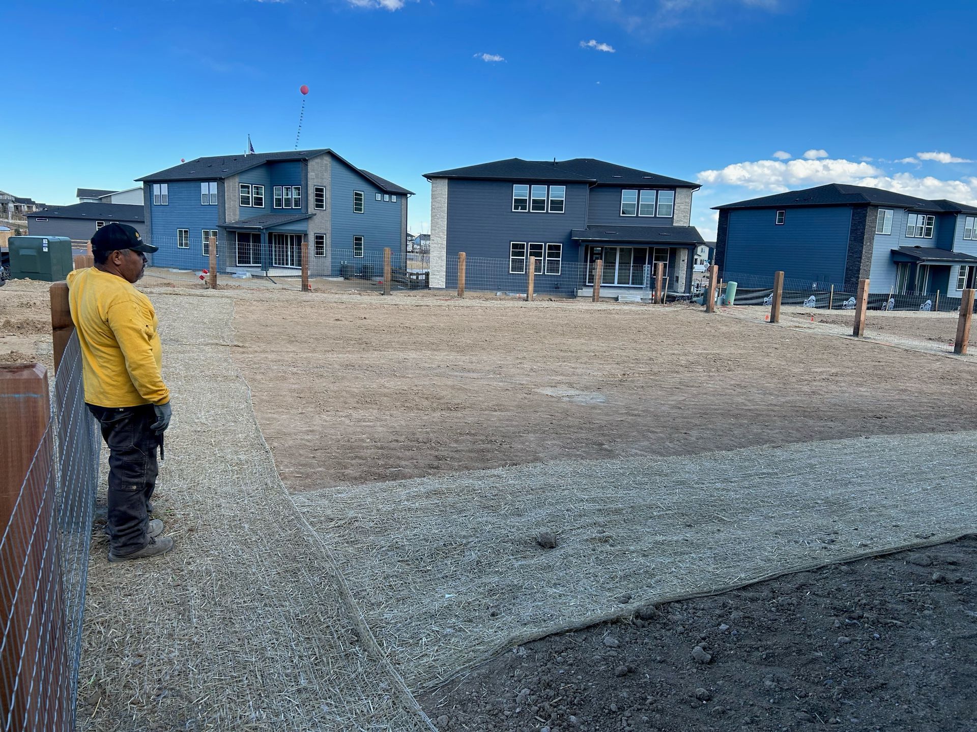 Person in a yellow jacket standing in a gravel lot with several blue houses in the background.