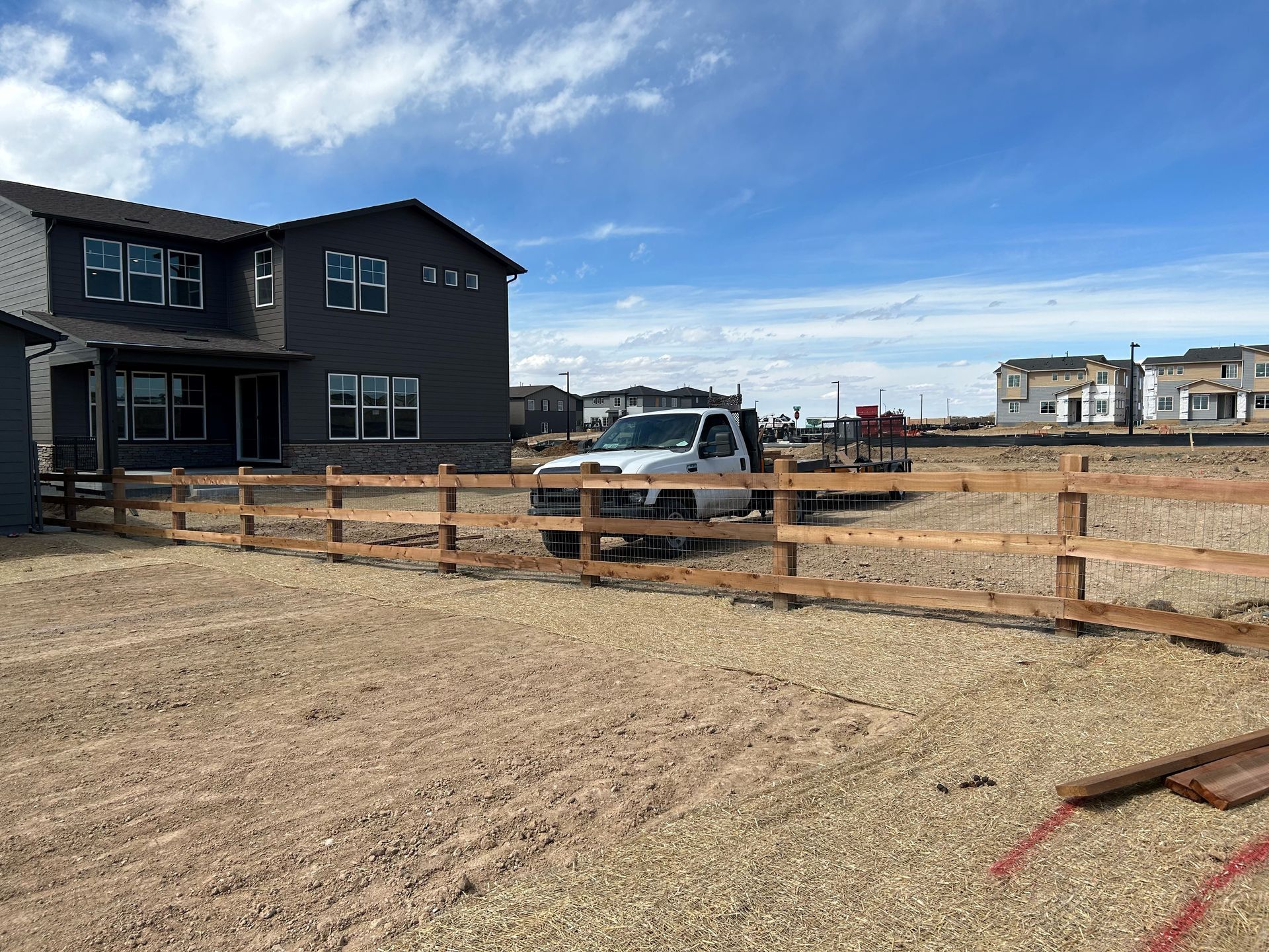 New houses under construction behind a wooden fence on a dirt lot under a blue sky
