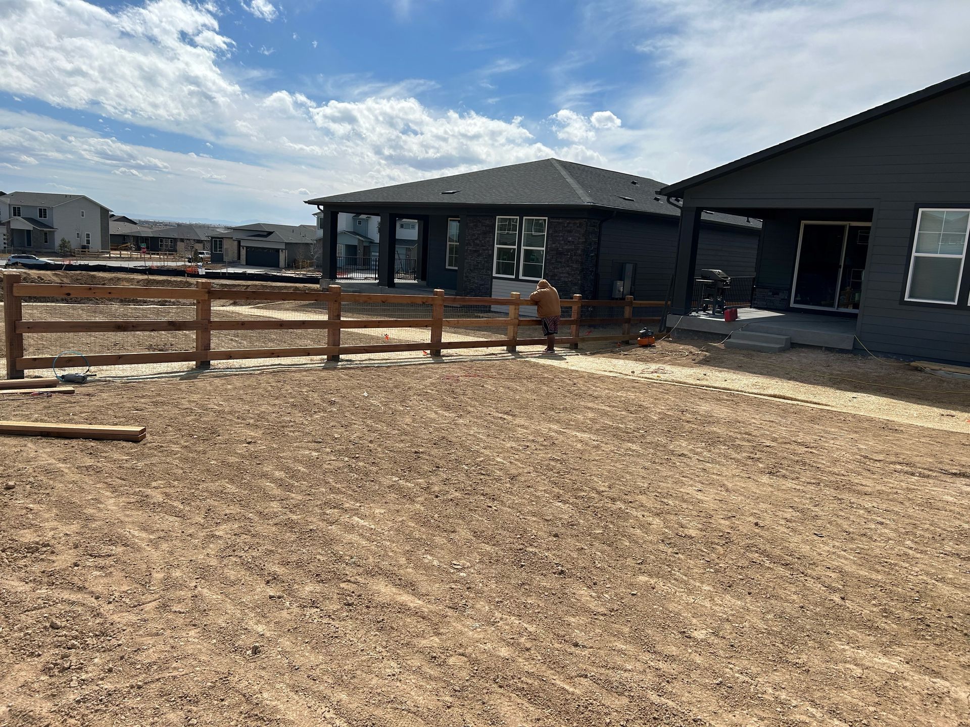 Two horses in a fenced dirt paddock beside dark gray buildings under a cloudy sky