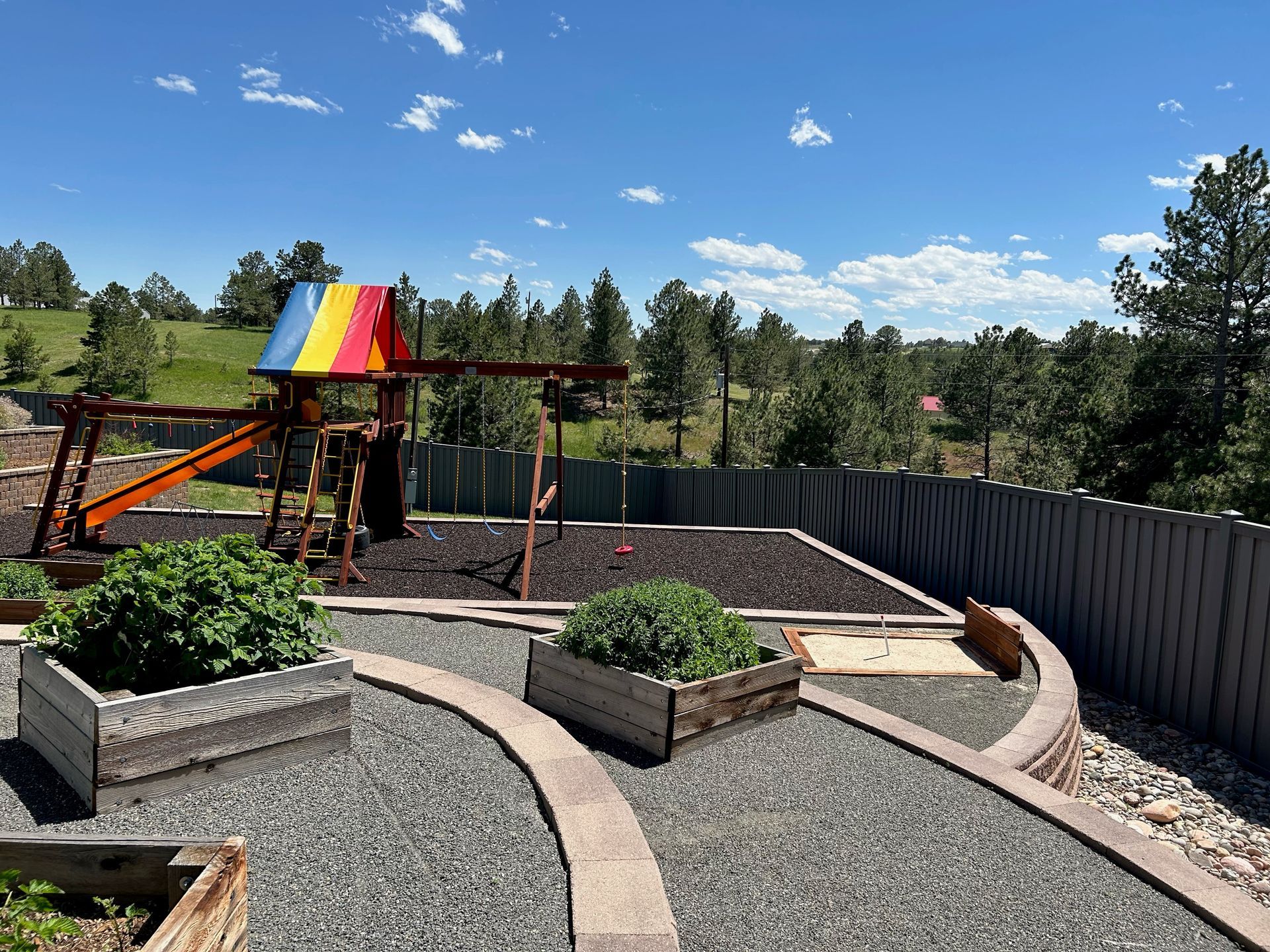 Backyard playground with rainbow roof, swings, gravel paths, raised planters, and a fenced yard under blue sky