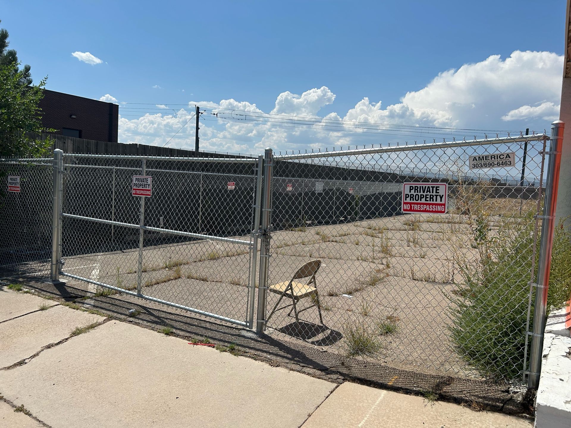 Chain-link gate blocking a sandy lot with a folding chair and red warning signs under a blue sky.