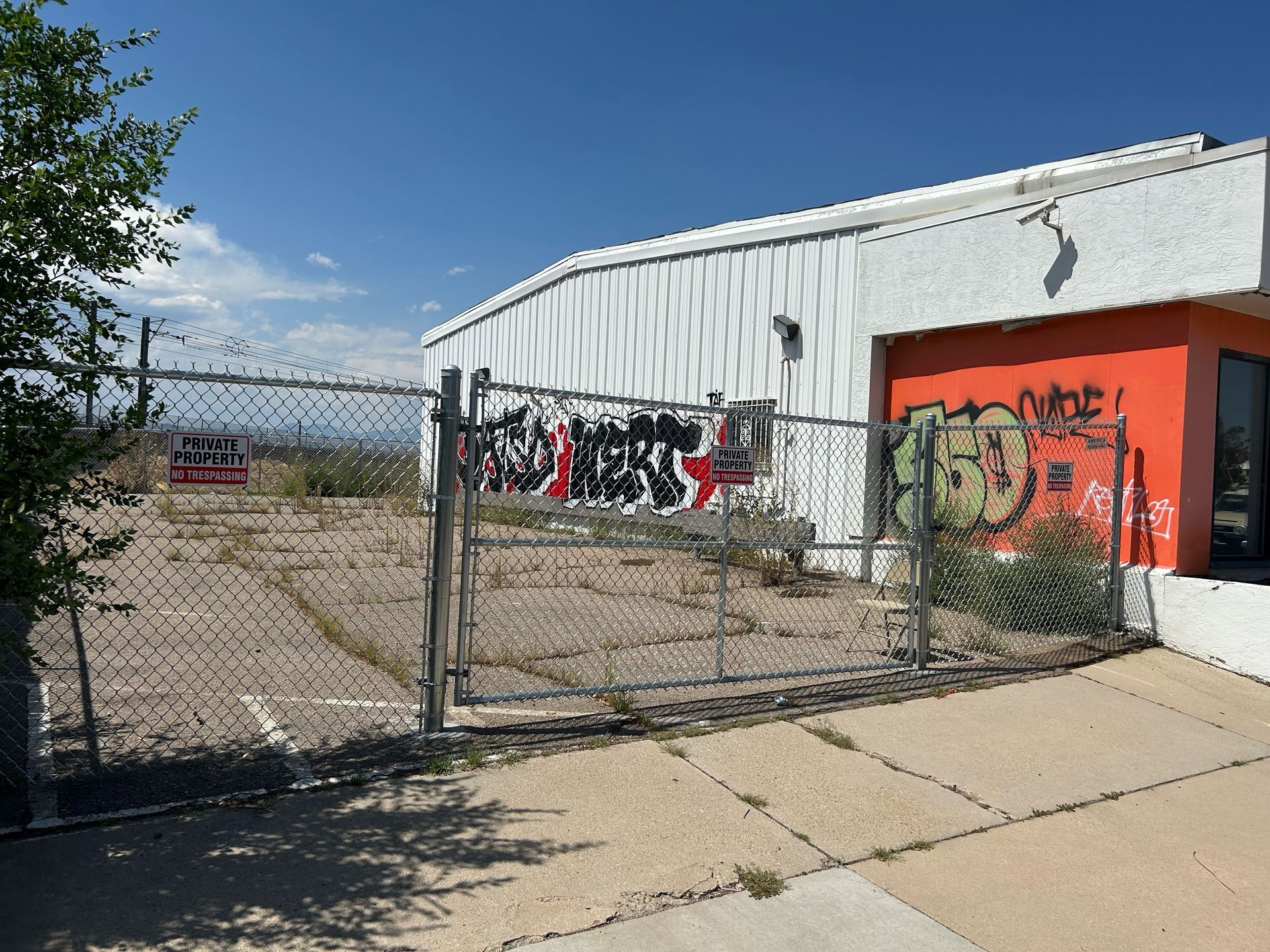 Chain-link gate beside a white building with orange door and graffiti, opening to a gravel lot