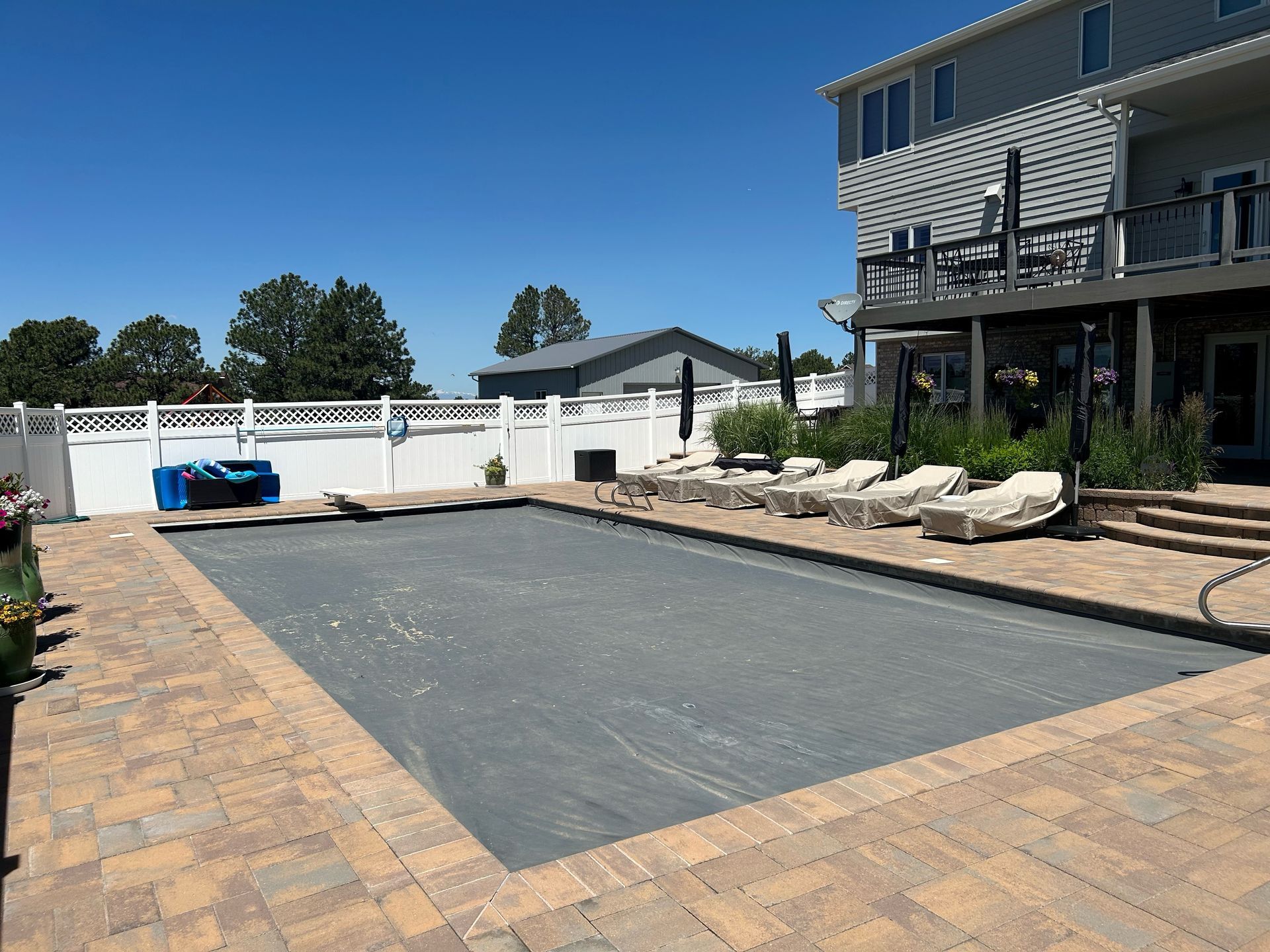 Empty rectangular backyard pool area with dark surface, white fence, lounge chairs, and two-story house.