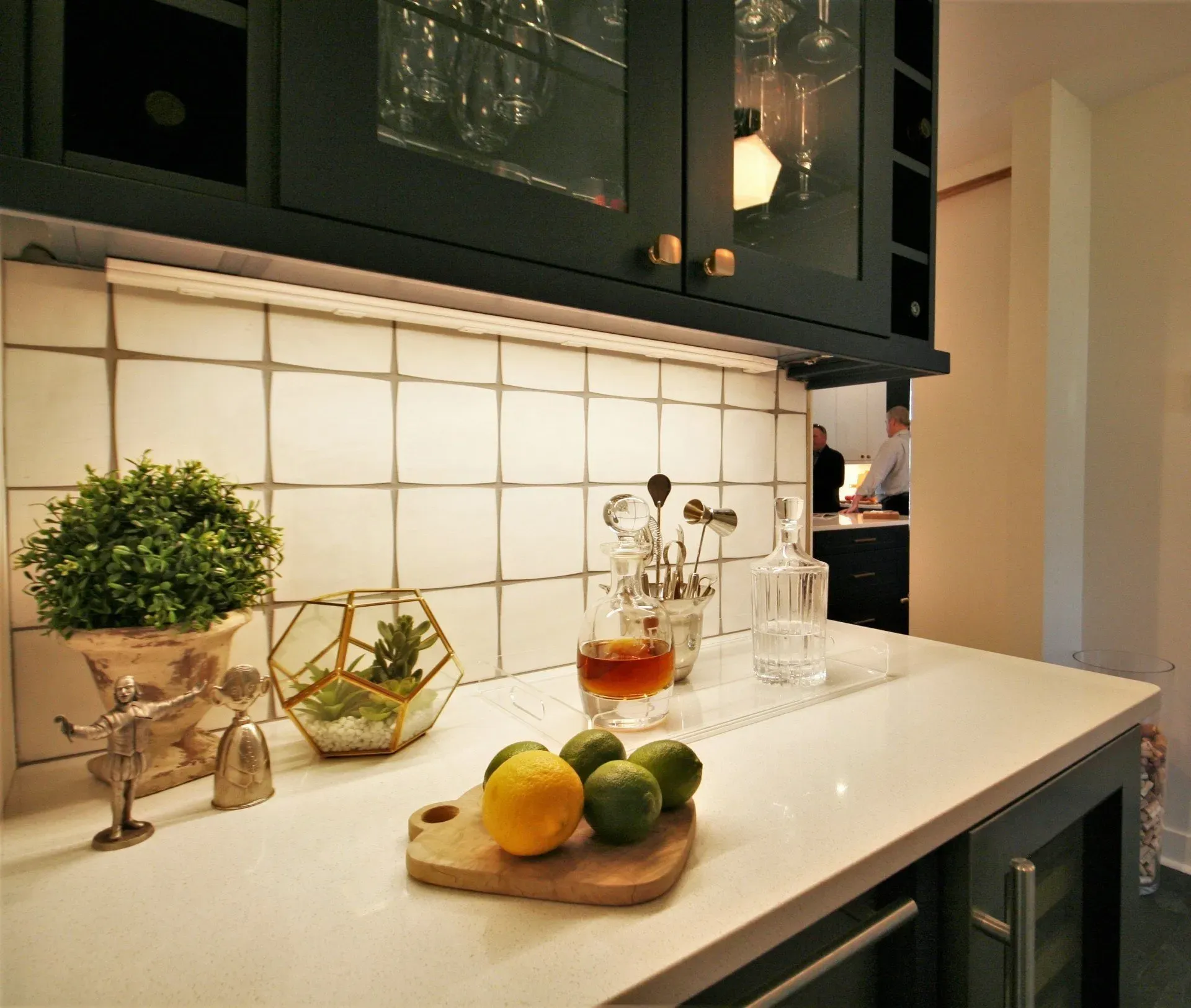 Kitchen counter with lemons, limes, drink pitcher, potted herb, and glassware under dark cabinets
