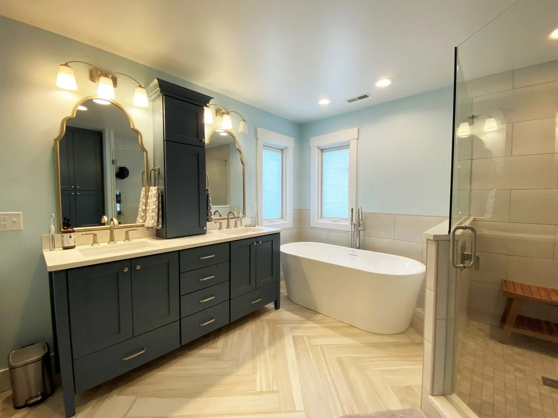 Modern bathroom with dark vanity, double mirrors, freestanding tub, and glass shower in a bright tiled room