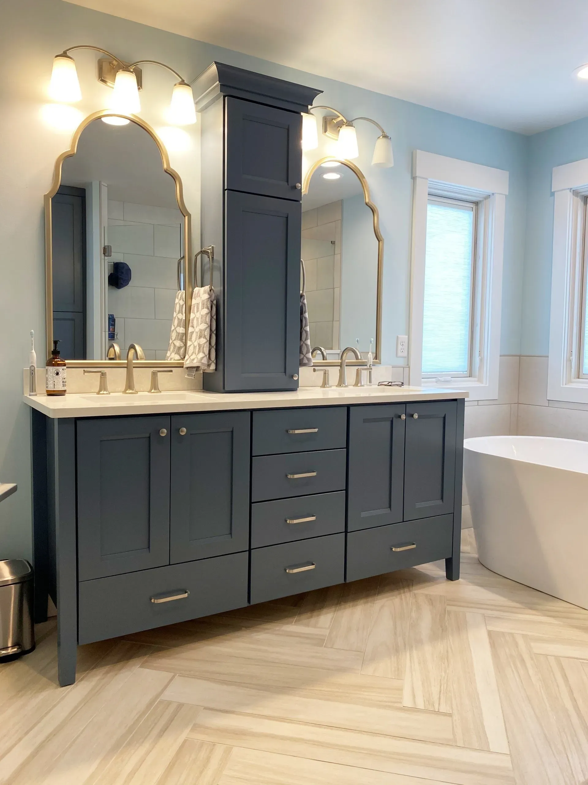 Blue bathroom vanity with twin gold mirrors, white counters, and a freestanding tub by a window.