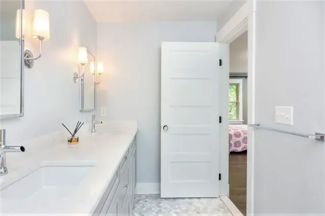 Bright white bathroom with double vanity, wall sconces, and open door to a bedroom with pink bedding