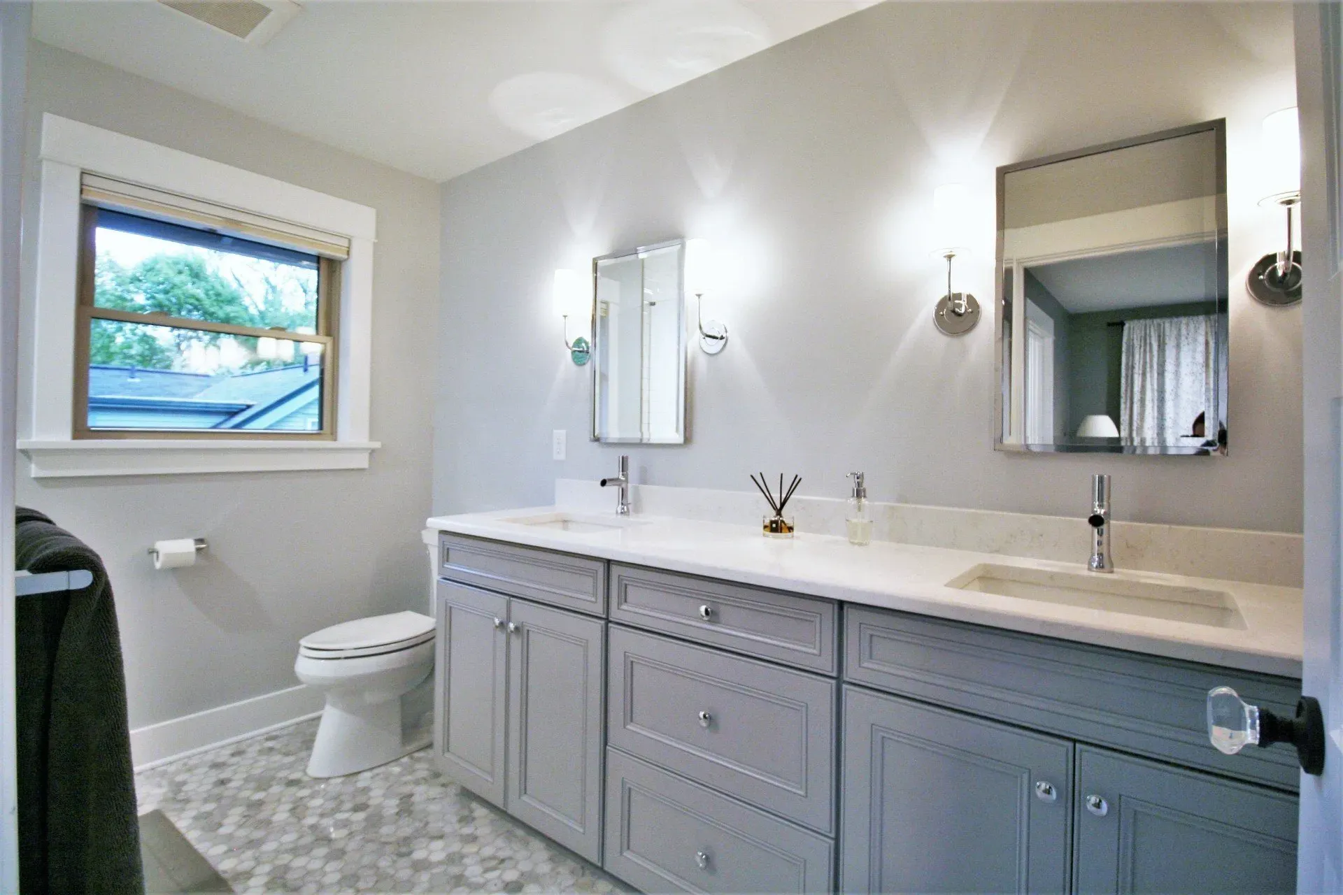 Bright gray bathroom with double vanity, mirror, toilet, and window overlooking greenery.