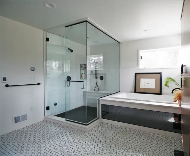Modern bathroom with glass shower, white walls, patterned tile floor, and bench by a small window.