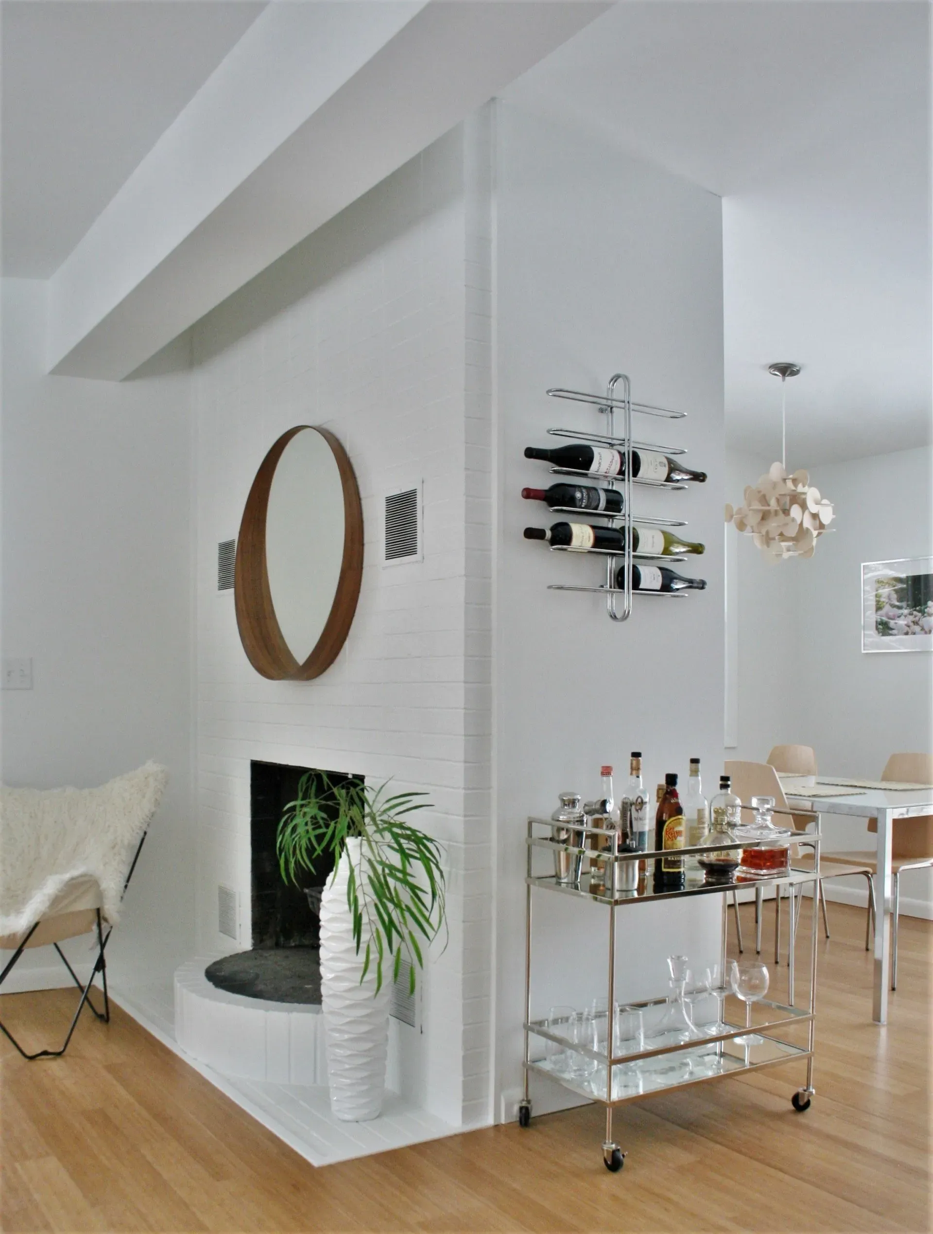 Modern open-plan dining area with white walls, wood floor, bar cart, wine rack, and round mirror.