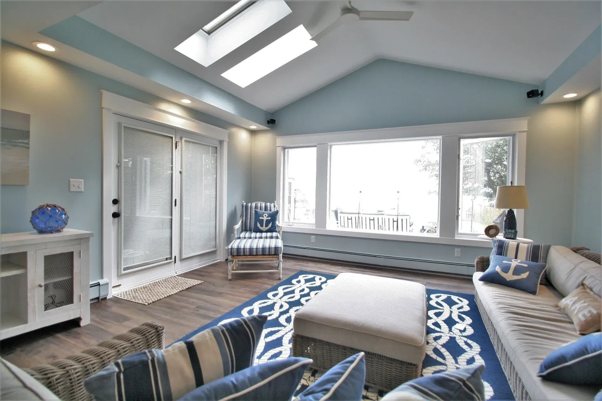 Bright living room with skylights, large window, blue-gray decor, and striped seating around a central ottoman
