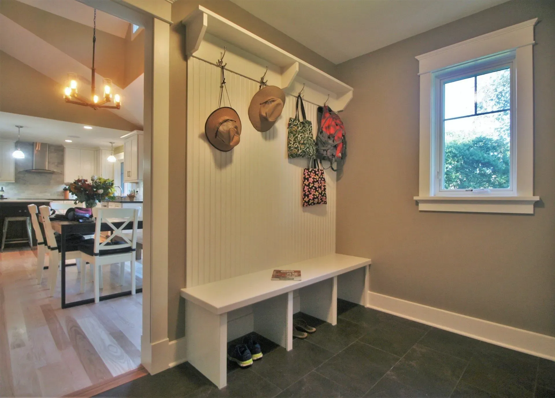 Small entryway bench with wall hooks, hats and bags, leading to a dining area and window.