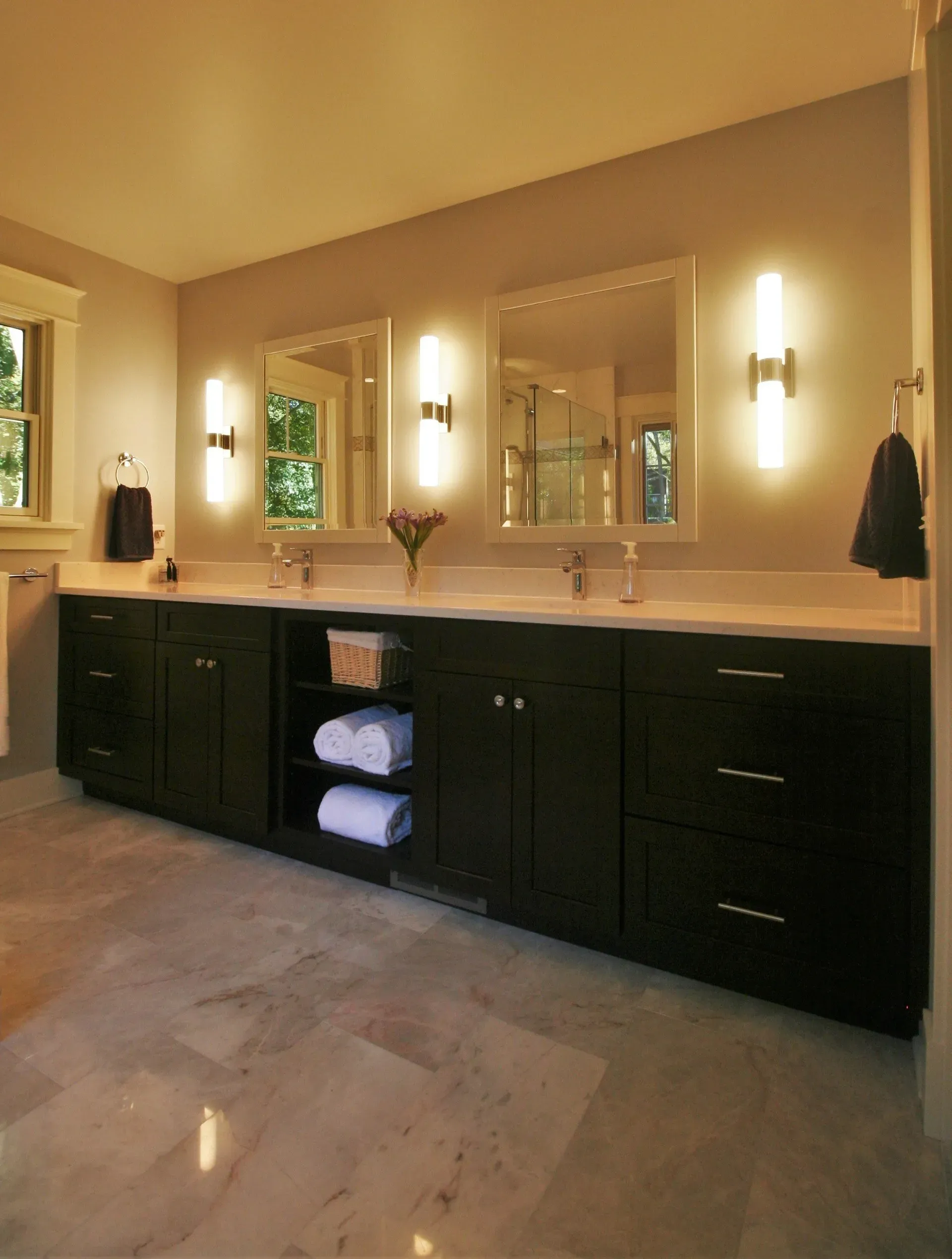 Elegant bathroom vanity with double sinks, wall sconces, large mirrors, and dark cabinetry under warm lighting
