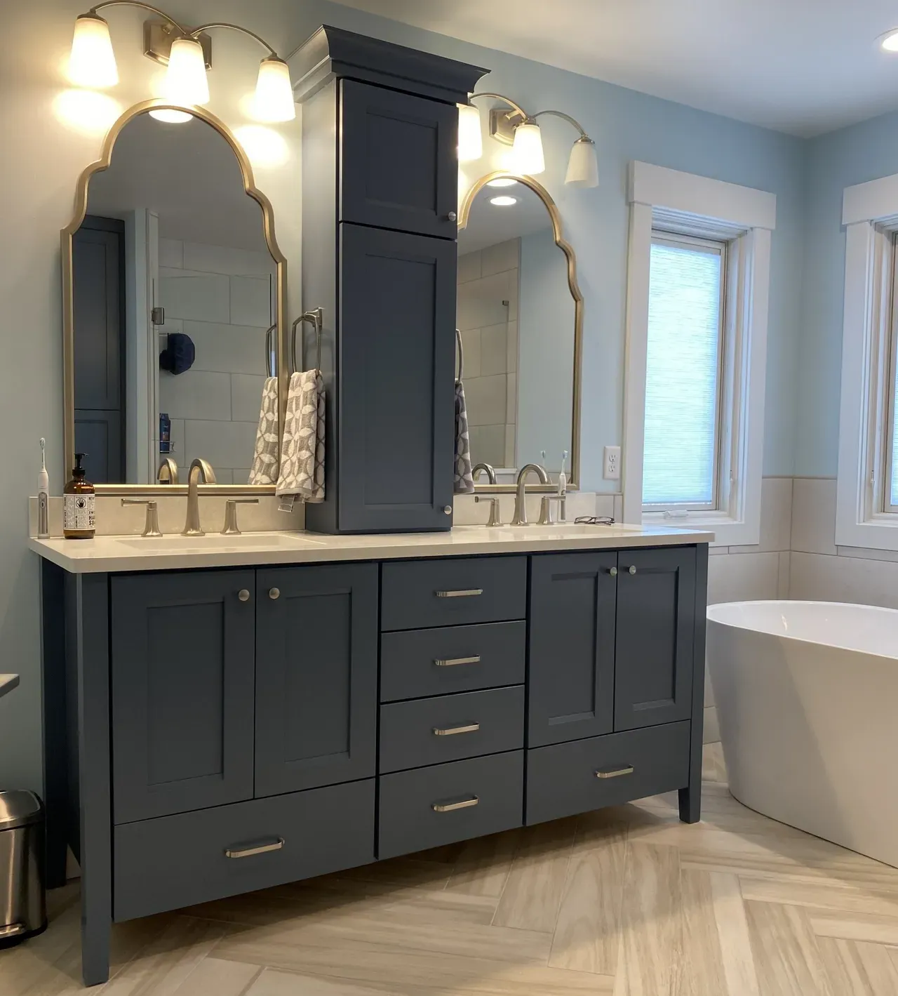 Dark blue double vanity with mirrors and sconces in a bright bathroom with a freestanding tub